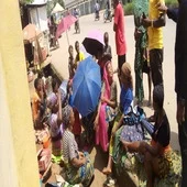 Beggars, including elderly men, women and children, seated along a busy Abuja street with bowls extended as pedestrians and vehicles pass by.