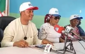 APC leaders seated at a press briefing table wearing party caps and addressing journalists